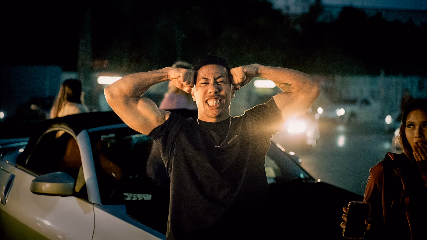 Man flexing his muscles and smiling while standing in front of a white car at night.